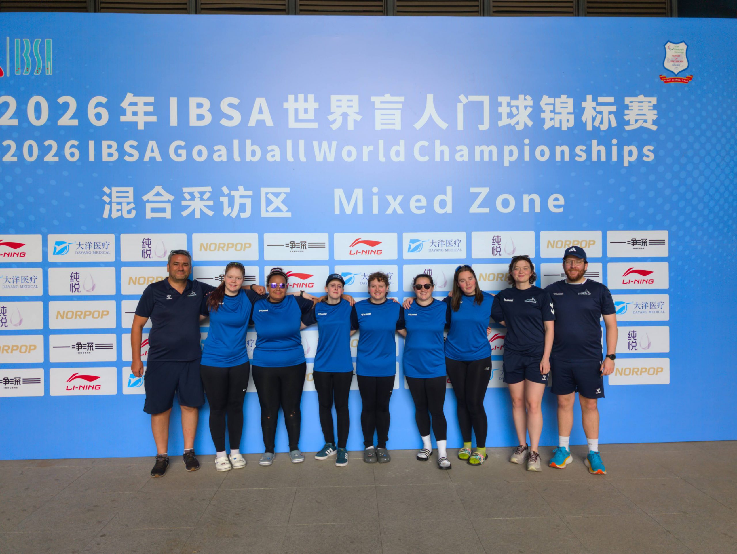 Group photo of the GB Women's squad and staff, stood in front of a large IBSA Goalball World Championships board.