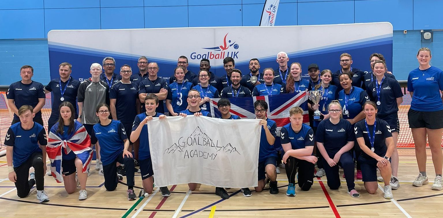 Photograph of the Great Britain Male and Female squads after winning the bronze medal at the Goalball UK International Trophy in July 2025, with the Goalball Academy down on one knee on the front row. Everyone is wearing matching navy blue shirts, some hold a white banner with "Goalball Academy" written, and a Goalball UK banner is visible in the background