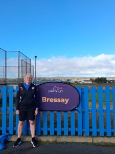 Kathryn stood next to a Bressay parkrun sign having just completed the 5k run. In the background, across the sea, houses in the town of Lerwick can be seen.